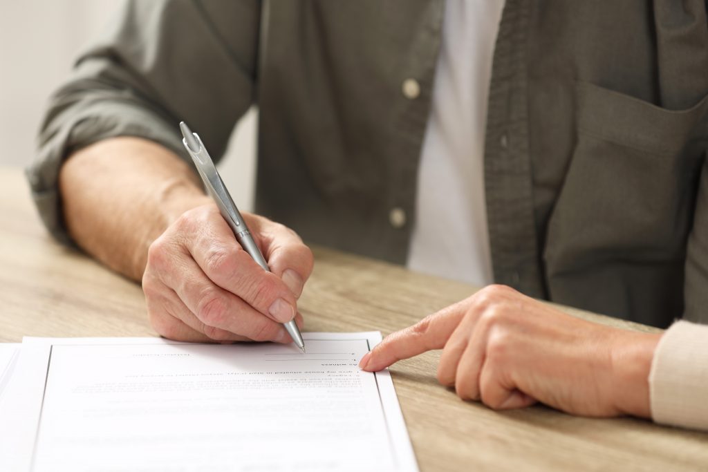 Notary pointing to senior man where to sign Last Will and Testament at wooden table indoors closeup - Guski ∙ Such - Rechtsanwälte Karlsruhe Vermächtnis