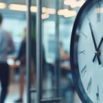 Close-up of a large analog clock with roman numerals; blurred office scene with people in background.