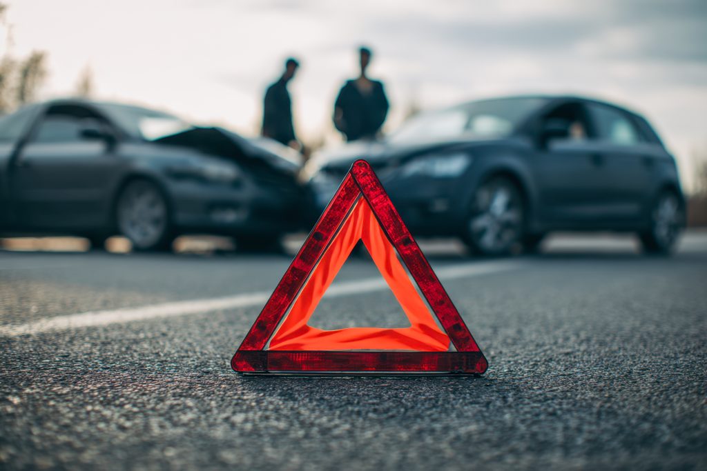 Reflective emergency warning triangle placed on road in front of two damaged vehicles with two people discussing car accident aftermath on cloudy day - Guski ∙ Such - Rechtsanwälte Karlsruhe Verkehrsunfall im Ausland: Wann gilt deutsches Recht? – LG Köln