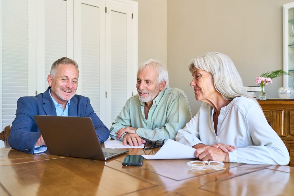 Retirement Planning And Financial Protection Senior Couple Receiving Professional Advice From Advisor On Laptop For Long-Term Security - Guski ∙ Such - Rechtsanwälte Karlsruhe Three seniors sit at a wooden table together, looking at a laptop and papers during a meeting.