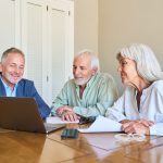 Three seniors sit at a wooden table together, looking at a laptop and papers during a meeting.