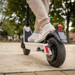 Person wearing beige pants and sneakers rides a white electric scooter; view focuses on rear wheel and brake light.