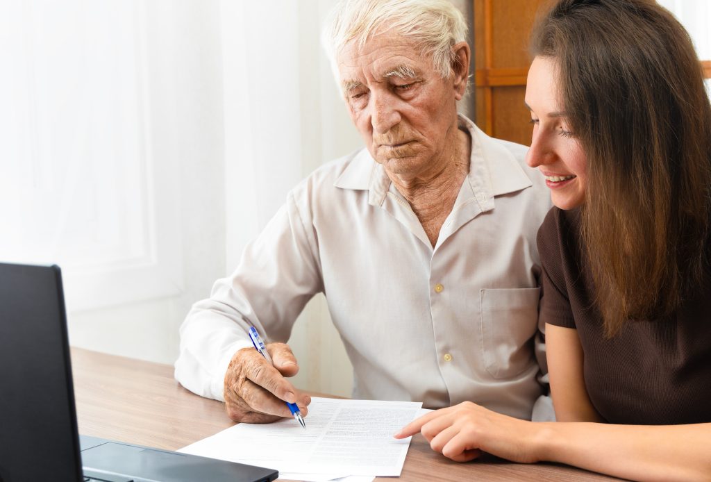 Older man and younger woman reviewing and signing documents at a desk with a laptop nearby.