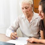 Older man and younger woman reviewing and signing documents at a desk with a laptop nearby.
