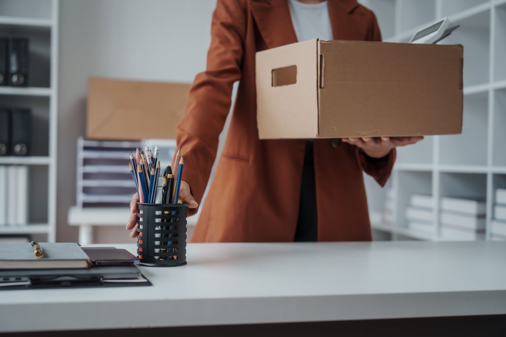 Kündigung vom Arbeitgeber oder selbst kündigen? Kündigungsarten, Fristen nach § 622 BGB, Schriftform und Kündigungsschutz verständlich erklärt. - Guski ∙ Such - Rechtsanwälte Karlsruhe Person in a brown blazer carries a cardboard box in a bright office space with a pencil cup on the desk in the foreground.