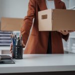 Person in a brown blazer carries a cardboard box in a bright office space with a pencil cup on the desk in the foreground.